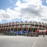 Nowoczesna chorten arena w Białymstoku, z charakterystyczną, ażurową konstrukcją dachu. Przed wejściem bramki obrotowe i łuk z logo stadionu.
