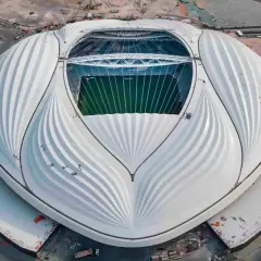 Al Janoub Stadium z lotu ptaka, z charakterystycznym białym dachem przypominającym żagiel. Widać zieloną murawę boiska i otaczające go tereny budowy.