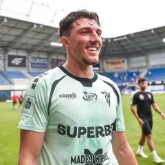 Uśmiechnięty Marcel Łubik w jasnej koszulce piłkarskiej na tle stadionu.