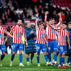 Piłkarze Atletico Madryt w pasiatych koszulkach świętują zwycięstwo. Wszyscy uśmiechnięci, niektórzy unoszą ręce w geście radości, kiedy gra Atletico Madryt przynosi sukces.