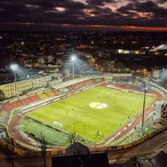 Wieczorny mecz na stadionie miejskim w Chojnicach. Piłkarze biegają po jasno oświetlonej murawie, a nad miastem zapada zmrok.