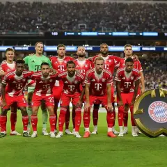 Drużyna Bayern Monachium – zawodnicy w czerwonych strojach pozują do zdjęcia na tle trybun. Obok nich stoi okrągłe logo klubu.