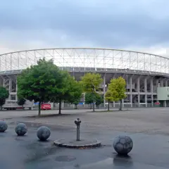 Ernst Happel Stadion z charakterystyczną białą kratownicą dachu, otoczony zielenią i betonowymi kulami przypominającymi piłki nożne.