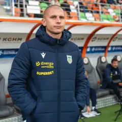 Trener Arki Gdynia, w kurtce z logo Energa i Superbet, stoi na tle ławki rezerwowych.