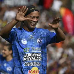 Uśmiechnięty Ronaldinho w niebieskiej koszulce z logo "FUTBOL CL" macha do kibiców. Widać, gdzie gra Ronaldinho.