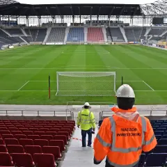 Pusta trybuna stadionu Pogoni Szczecin, z dwoma pracownikami w kamizelkach odblaskowych.