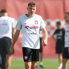 Szymon Żurkowski w białej koszulce reprezentacji Polski z logo Orlen i InPost, na tle czerwonej bandy.