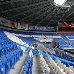Puste trybuny Cardiff City Stadium, z niebieskimi i białymi siedzeniami, ozdobione banerem "Visit Malaysia".