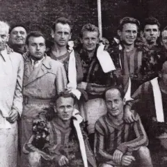 Drużyna piłkarska z wieńcami laurowymi, być może świętująca zwycięstwo. W tle widać fragmenty strojów sportowych, a jeden z mężczyzn ma na sobie płaszcz.