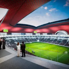 Widok na zieloną murawę stadionu Al Bayt, otoczoną trybunami pełnymi widzów. Czerwony dach tworzy imponującą konstrukcję.