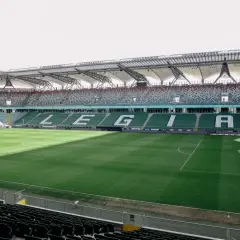 Puste trybuny stadionu Wojska Polskiego czekają na kibiców. Na zielonej murawie widać białe linie boiska.