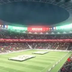 Widok na wypełniony po brzegi, największy stadion na świecie. Na murawie rozłożone wielkie transparenty, a trybuny rozświetlone flagami i światłami.
