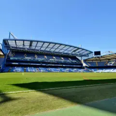 Pusta trybuna nowego stadionu Chelsea, z zieloną murawą i niebieskimi siedzeniami.