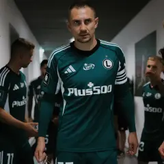 Znani polscy piłkarze w zielonych strojach z białymi pasami na ramionach, idący korytarzem.