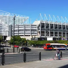 St James' Park w Newcastle, z charakterystyczną białą konstrukcją dachu, otoczony zielenią i ruchliwą ulicą z autobusem i rowerzystą.