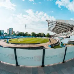 Stadion miejski w Rzeszowie z zieloną murawą, bieżnią żużlową i niebieskimi trybunami. W tle budynki mieszkalne i błękitne niebo z chmurami.