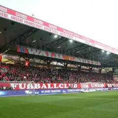 Trybuna na stadionie Union Berlin wypełniona kibicami. Na czerwonym banerze widnieje napis "UNSERE LIEBE. UNSERE MANNSCHAFT. UNSER STOLZ. UNSER VEREIN.".