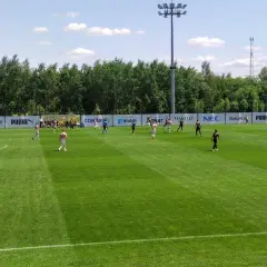 Piłkarze grają na boisku podczas meczu Betclic 3 liga. Słońce świeci, a drzewa tworzą tło.