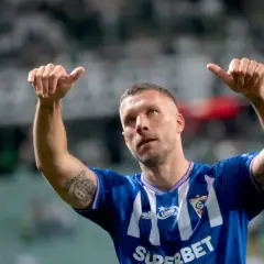 Piłkarz Louis Gabriel Podolski w niebieskiej koszulce z białymi pasami, z uniesionymi kciukami, świętuje zwycięstwo na boisku.