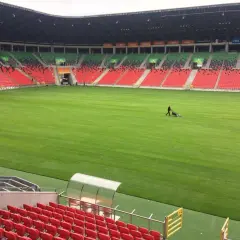 Pracownicy koszą trawę na boisku stadionu GKS Tychy. Puste trybuny w kolorach czerwonym i zielonym czekają na kibiców.