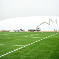 Budowa hali pneumatycznej na stadionie Ślęzy Wrocław. Sztuczna murawa z białymi liniami, koparka i gruszka do betonu w tle.
