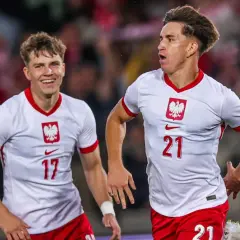Dwóch młodych zawodników w białych koszulkach z orłem na piersi, z numerami 17 i 21, cieszy się po zdobytej bramce.