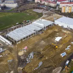 Budowa obiektu sportowego w Radzionkowie. Widać plac budowy z fundamentami, konstrukcją hali, maszynami budowlanymi i pobliski stadion.