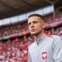 Sebastian Szymański w szarej bluzie z orłem na piersi, na tle trybun wypełnionych kibicami w biało-czerwonych barwach.