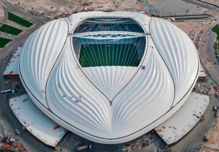 Al Janoub Stadium z lotu ptaka, z charakterystycznym białym dachem przypominającym żagiel. Widać zieloną murawę boiska i otaczające go tereny budowy.