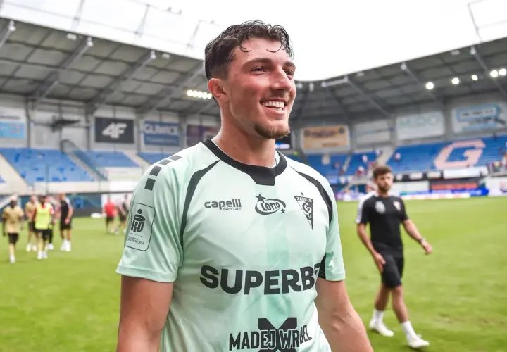 Uśmiechnięty Marcel Łubik w jasnej koszulce piłkarskiej na tle stadionu.
