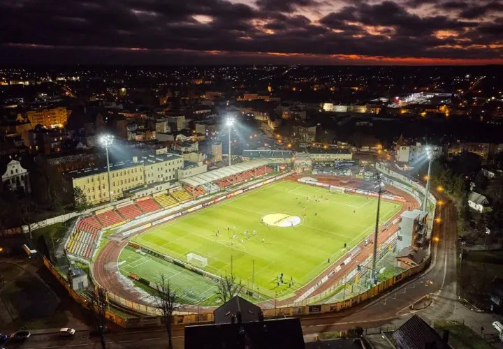 Wieczorny mecz na stadionie miejskim w Chojnicach. Piłkarze biegają po jasno oświetlonej murawie, a nad miastem zapada zmrok.