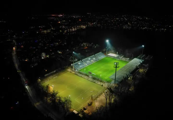 Nocne oświetlenie rozświetla zieloną murawę stadionu Sokoła w Ostródzie, otoczonego ciemnością i miejskimi światłami.