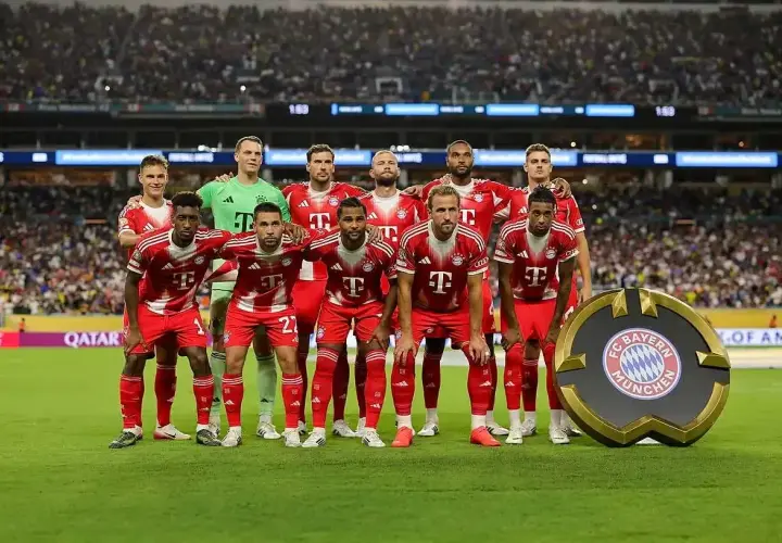 Drużyna Bayern Monachium – zawodnicy w czerwonych strojach pozują do zdjęcia na tle trybun. Obok nich stoi okrągłe logo klubu.