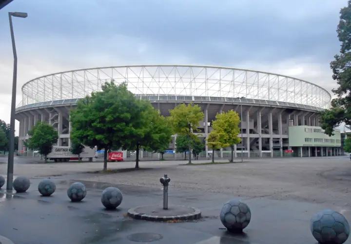Ernst Happel Stadion z charakterystyczną białą kratownicą dachu, otoczony zielenią i betonowymi kulami przypominającymi piłki nożne.