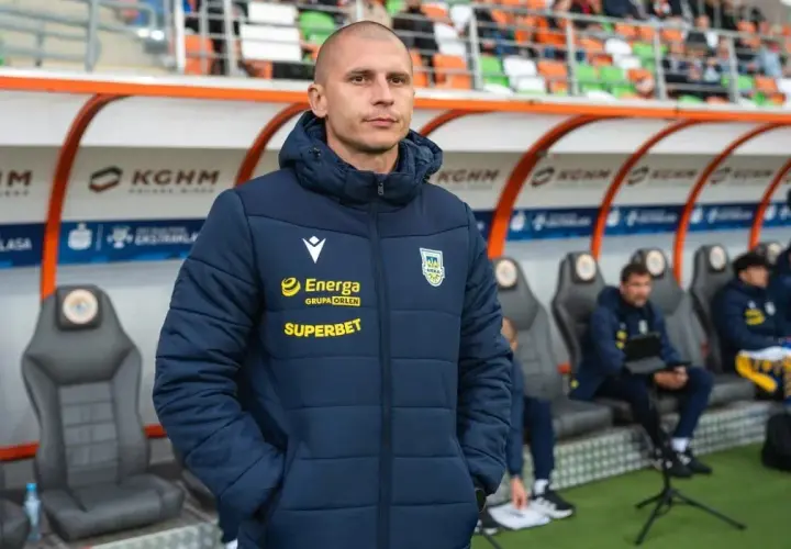 Trener Arki Gdynia, w kurtce z logo Energa i Superbet, stoi na tle ławki rezerwowych.