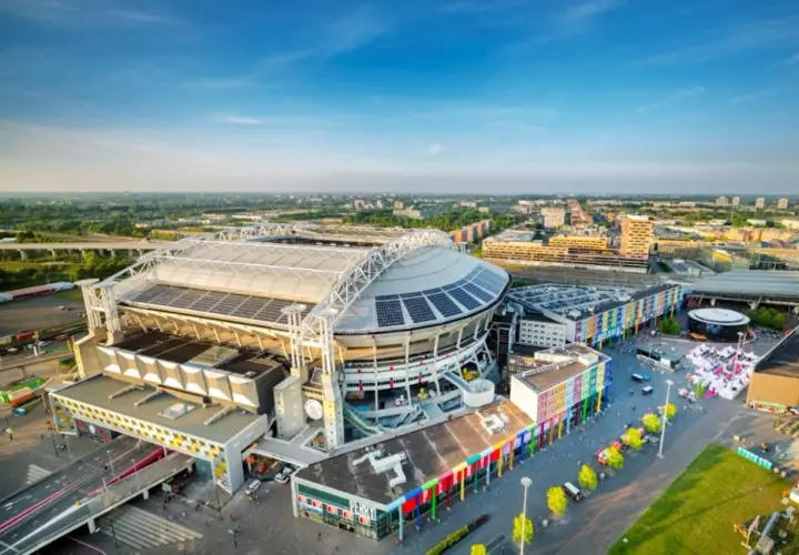 Johan Cruyff Arena, imponujący stadion z panelami słonecznymi na dachu, otoczony kolorowymi budynkami i zielenią.