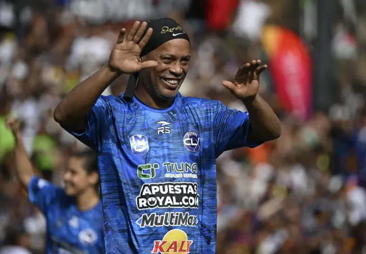 Uśmiechnięty Ronaldinho w niebieskiej koszulce z logo "FUTBOL CL" macha do kibiców. Widać, gdzie gra Ronaldinho.