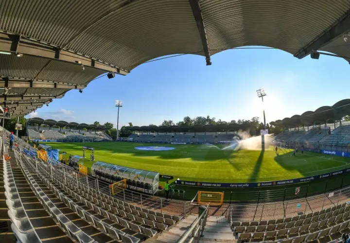 Pusta trybuna stadionu Miedzi Legnica, zielona murawa i zraszacze tworzą spokojny obraz przed meczem.