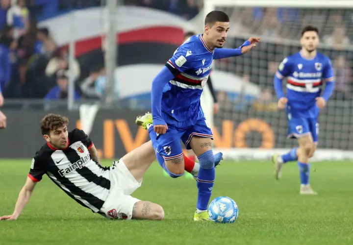 Piłkarze w akcji podczas rozgrywek Serie B. Jeden z zawodników w niebieskim stroju próbuje odebrać piłkę leżącemu na trawie przeciwnikowi w czarno-białej koszulce.