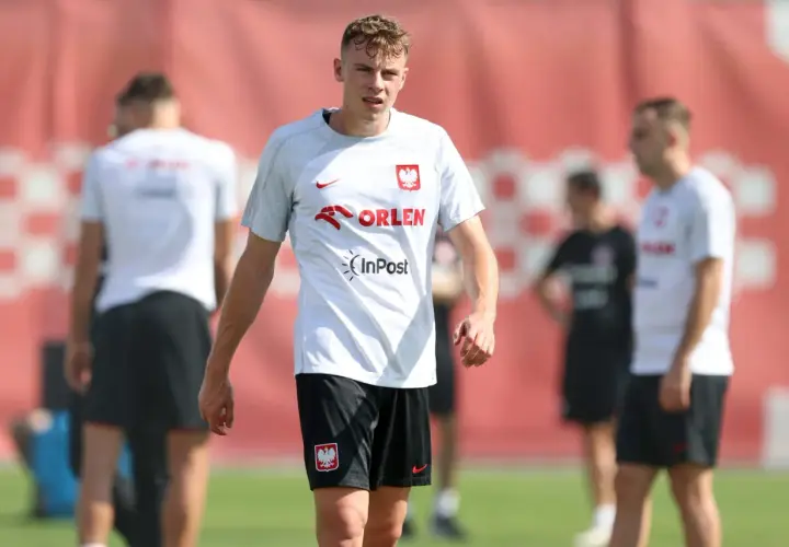 Szymon Żurkowski w białej koszulce reprezentacji Polski z logo Orlen i InPost, na tle czerwonej bandy.