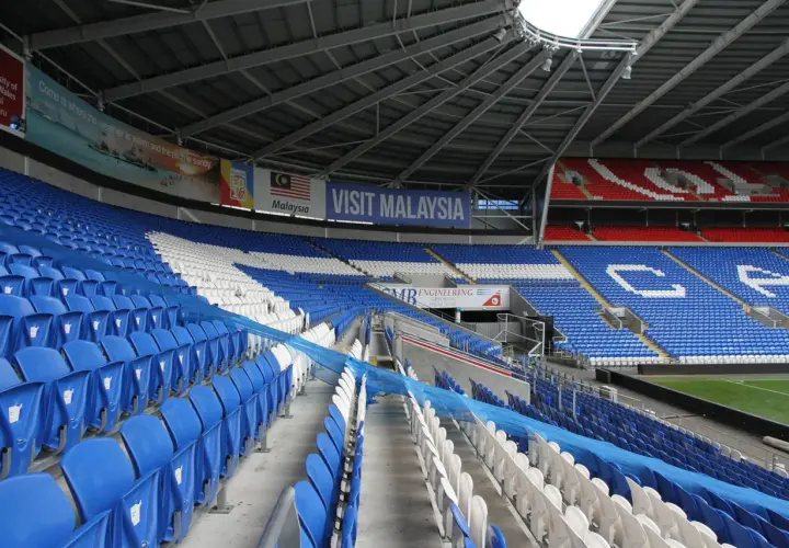 Puste trybuny Cardiff City Stadium, z niebieskimi i białymi siedzeniami, ozdobione banerem "Visit Malaysia".