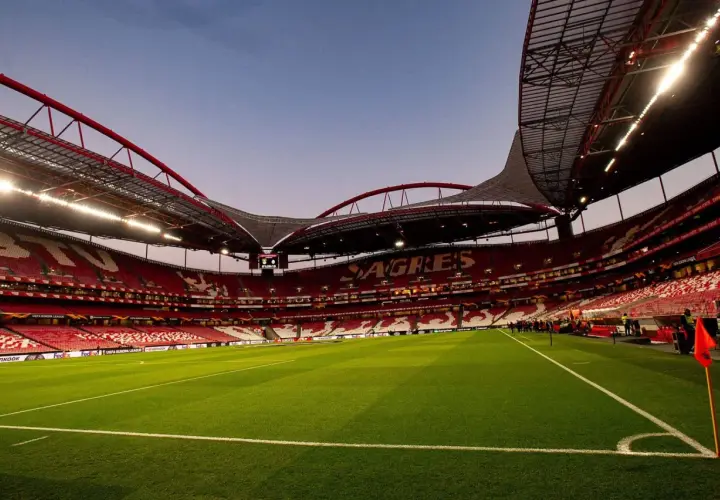 Pusta trybuna stadionu **Estadio da Luz** w Lizbonie, z zieloną murawą i czerwonymi siedzeniami.