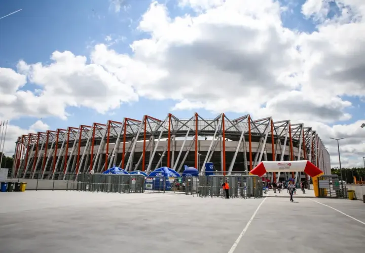 Nowoczesna chorten arena w Białymstoku, z charakterystyczną, ażurową konstrukcją dachu. Przed wejściem bramki obrotowe i łuk z logo stadionu.