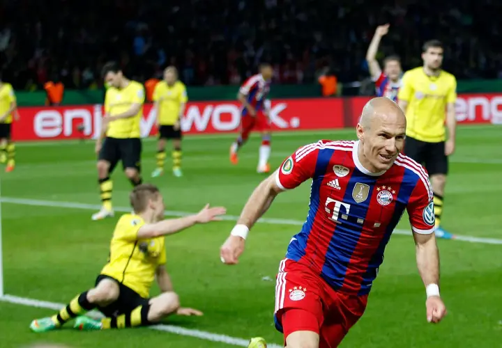 Piłkarz Bayernu w czerwono-niebieskiej koszulce cieszy się z bramki. W tle zawodnicy Borussii Dortmund w żółtych strojach.