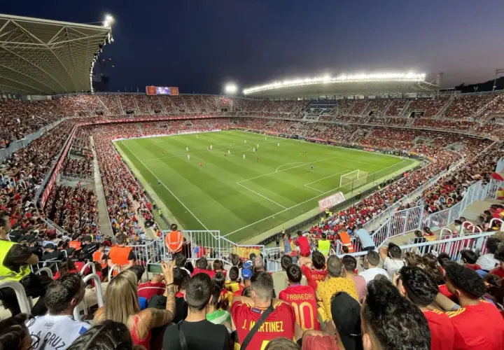 Widok z trybun na pełny stadion **Estadio La Rosaleda** podczas meczu piłki nożnej. Tłum kibiców w czerwonych barwach wypełnia trybuny.