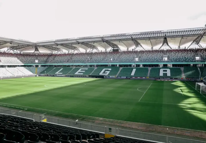 Puste trybuny stadionu Wojska Polskiego czekają na kibiców. Na zielonej murawie widać białe linie boiska.