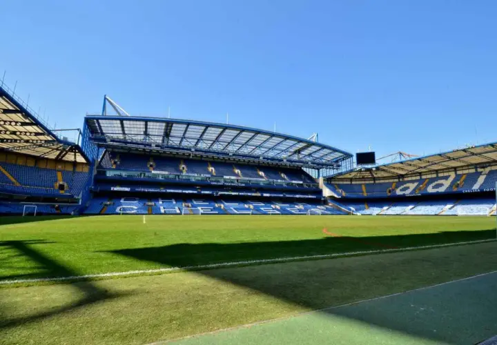 Pusta trybuna nowego stadionu Chelsea, z zieloną murawą i niebieskimi siedzeniami.