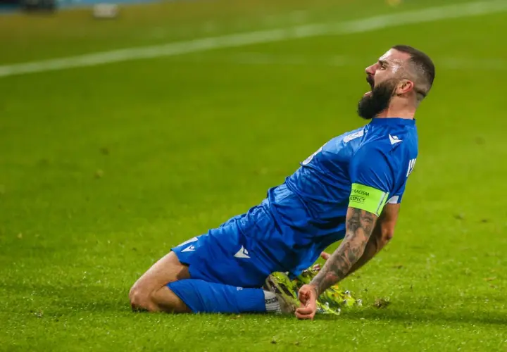 Mikael Ishak świętuje bramkę, klęcząc na zielonej murawie.