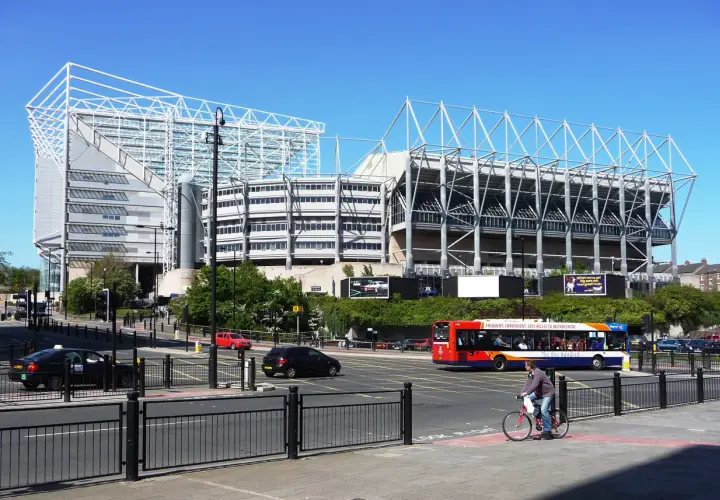 St James' Park w Newcastle, z charakterystyczną białą konstrukcją dachu, otoczony zielenią i ruchliwą ulicą z autobusem i rowerzystą.
