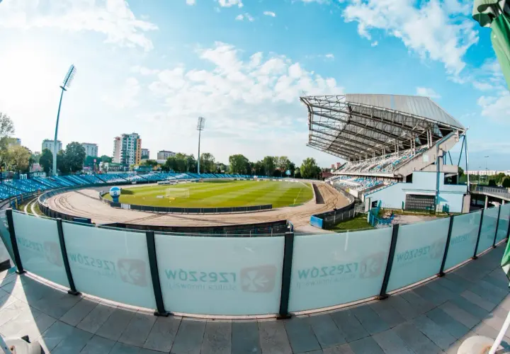 Stadion miejski w Rzeszowie z zieloną murawą, bieżnią żużlową i niebieskimi trybunami. W tle budynki mieszkalne i błękitne niebo z chmurami.